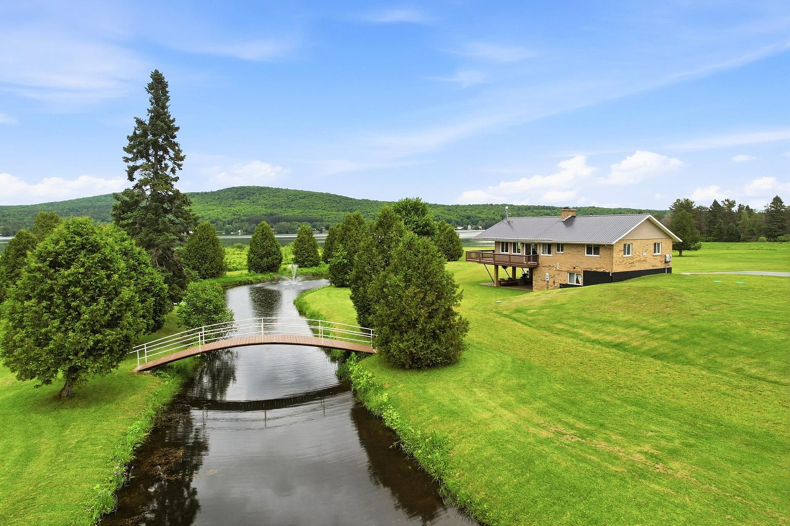 This elevated aerial perspective showcases a charming brick home situated on a sprawling, lush green lawn. The property features a picturesque stream crossed by a wooden arched bridge, leading toward a serene pond with a fountain, all set against a backdrop of rolling hills and a distant lake. The home includes a prominent wooden deck overlooking the grounds, emphasizing a peaceful, rural lifestyle.