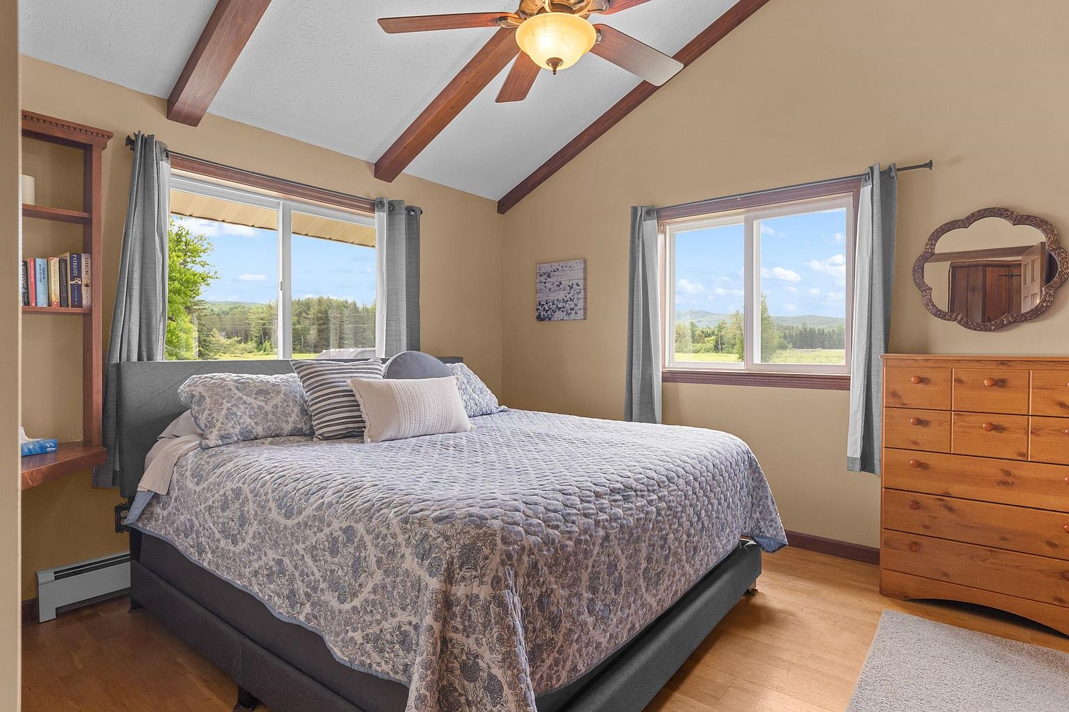 This primary bedroom features a vaulted ceiling with exposed wooden beams and a central ceiling fan. A large bed with a patterned blue and white quilt is positioned between two windows that offer views of the surrounding landscape. To the right, a wooden dresser is topped with a decorative mirror, while a built-in shelving unit on the left provides storage for books.