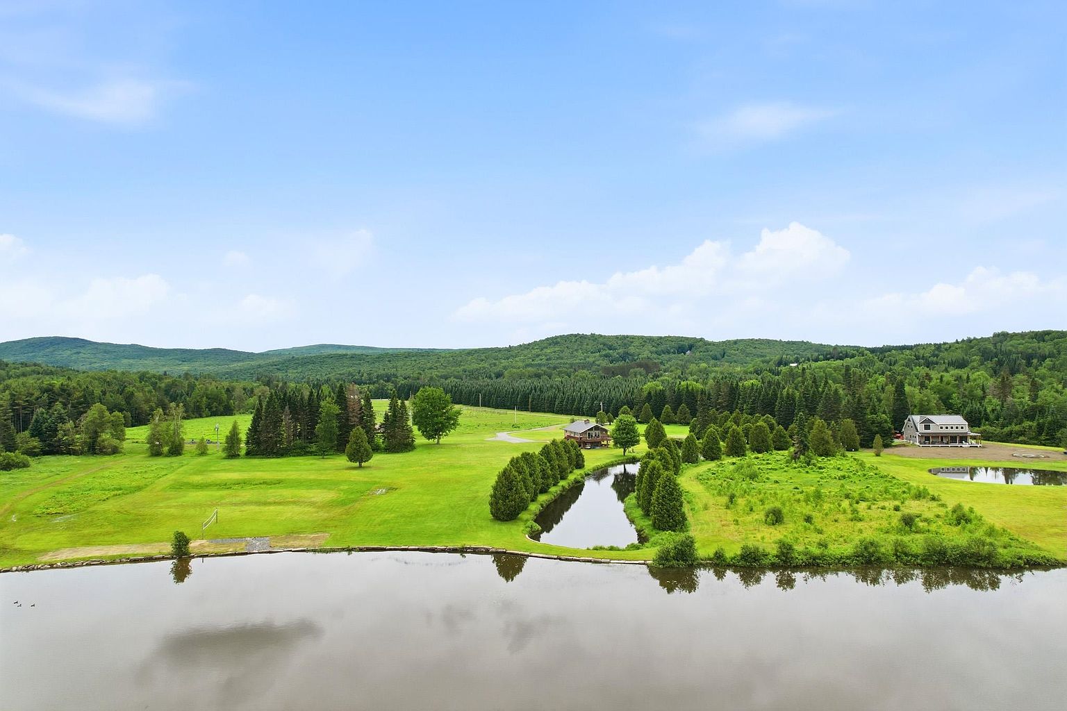 This expansive aerial view captures a sprawling rural property featuring a large pond in the foreground and a secondary smaller pond near a multi-story house. The landscape is characterized by lush green lawns, mature evergreen trees, and a dense forest backdrop under a bright, clear sky. The perspective highlights the vastness of the grounds and the peaceful, secluded setting of the residence.