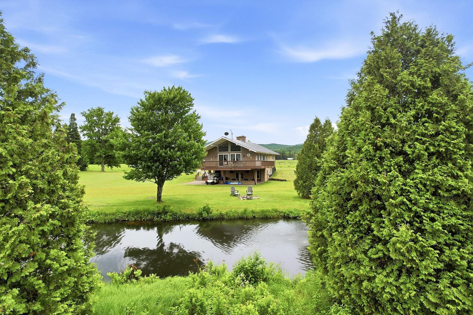This rear view of a multi-level home showcases a spacious wooden deck overlooking a lush green lawn and a small pond in the foreground. The house features a prominent A-frame style roofline with large windows that capture natural light, set against a backdrop of rolling hills and a clear blue sky. The property is framed by mature evergreen trees, creating a serene and private atmosphere.