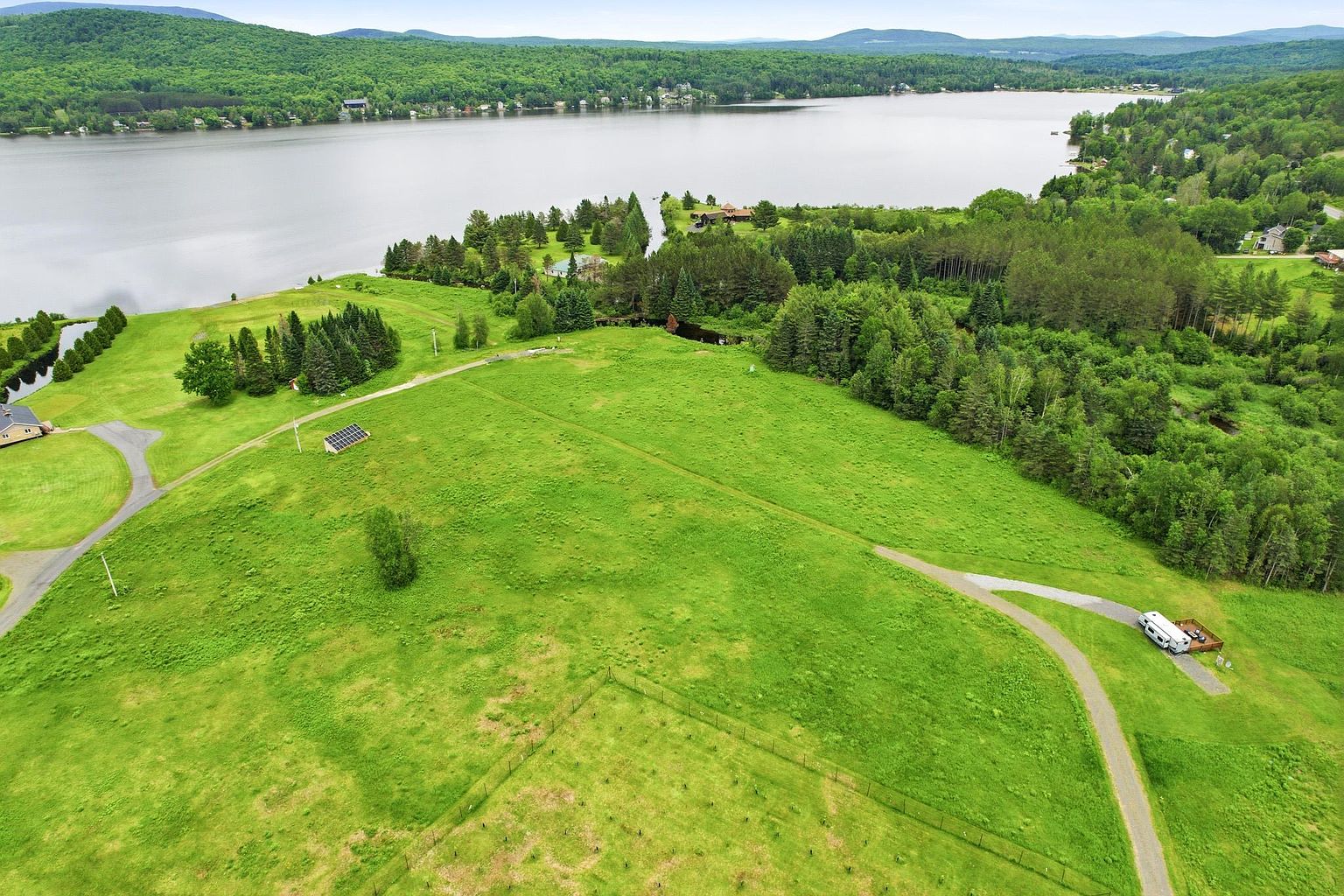 This expansive aerial view captures a large, grassy property sloping gently toward a serene lake surrounded by lush, dense forest. The landscape features a winding gravel driveway, a small solar panel array, and a recreational vehicle parked on a wooden deck, emphasizing the property's potential for outdoor living and development. The perspective provides a comprehensive look at the natural topography and the peaceful, secluded setting of the land.