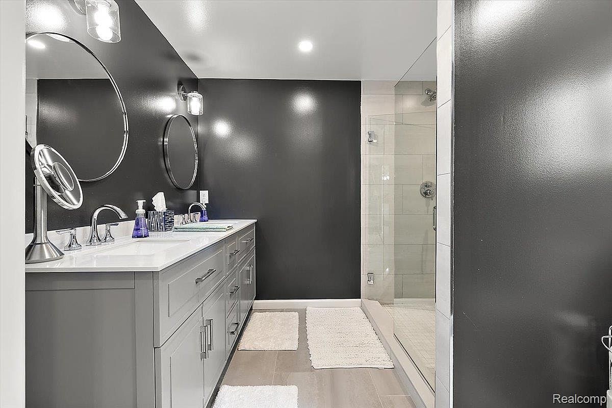 This is a primary bathroom featuring a double vanity with gray cabinets and a white countertop. The walls are painted a dark gray, and there are round mirrors above each sink. A glass-enclosed shower is visible to the right, and white rugs are placed on the gray tile floor. The overall impression is modern and stylish.