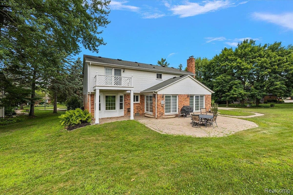 This is a rear view of a two-story house with a brick and white siding exterior. The house features a second-story balcony, a patio with outdoor furniture, and a well-maintained lawn. Mature trees surround the property, creating a sense of privacy and tranquility.
