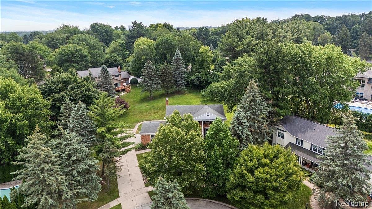 This aerial view showcases a well-maintained property nestled among lush greenery. Several houses are visible, each with unique architectural styles and surrounded by mature trees. A swimming pool can be seen in the background, suggesting a community or private amenity.