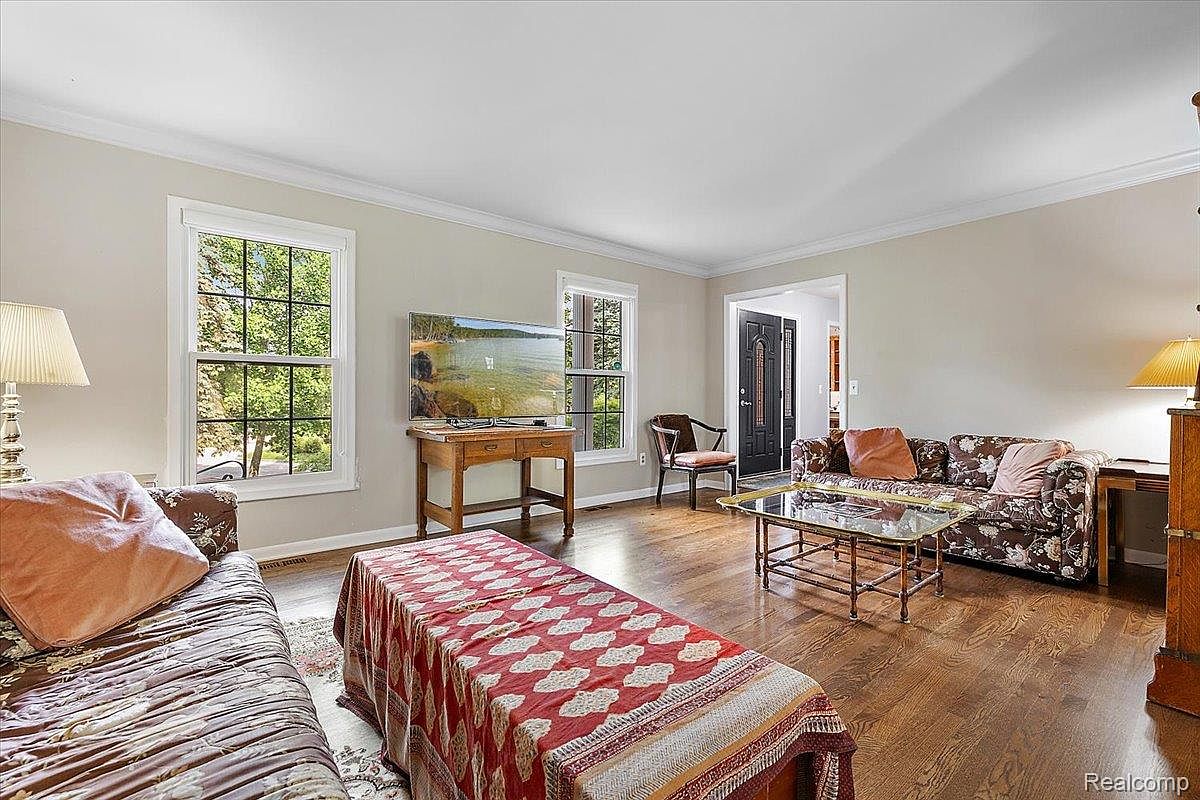 This is an interior shot of a living room featuring hardwood floors, neutral-colored walls, and a mix of traditional and eclectic furniture. The room has two large windows providing natural light, and a television is mounted above a wooden console table. The space is furnished with two sofas, a glass-topped coffee table, and a decorative trunk, creating a comfortable and inviting atmosphere.