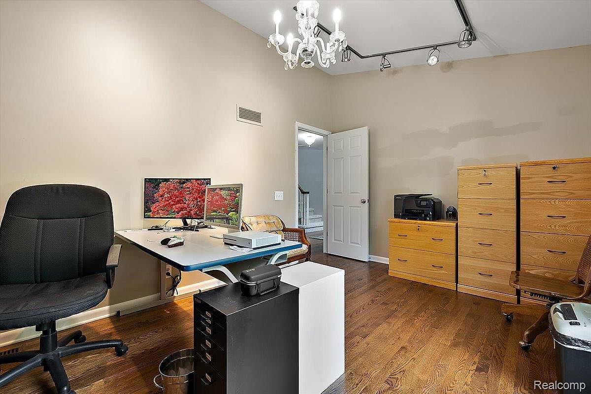 This is an interior shot of a home office. The room features hardwood floors, beige walls, and a white ceiling with a chandelier and track lighting. There is a desk with computer monitors, a black office chair, filing cabinets, and a door leading to another room, creating a functional and organized workspace.