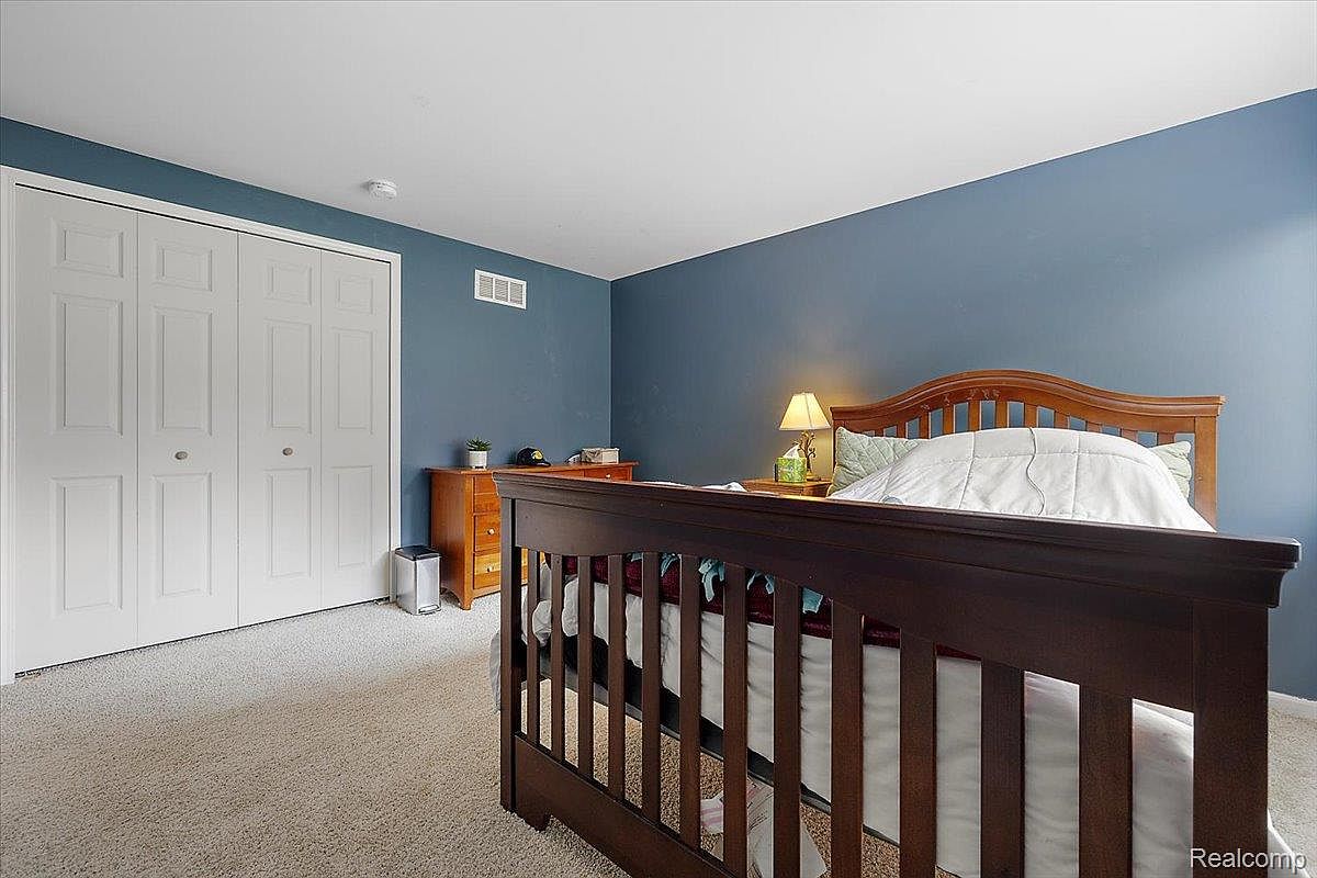 This is an interior shot of a bedroom featuring a dark wood crib in the foreground, a bed with white linens, and a blue accent wall. A wooden dresser and a white closet are visible in the background, adding to the room's functionality. The room is carpeted, creating a cozy and inviting atmosphere.