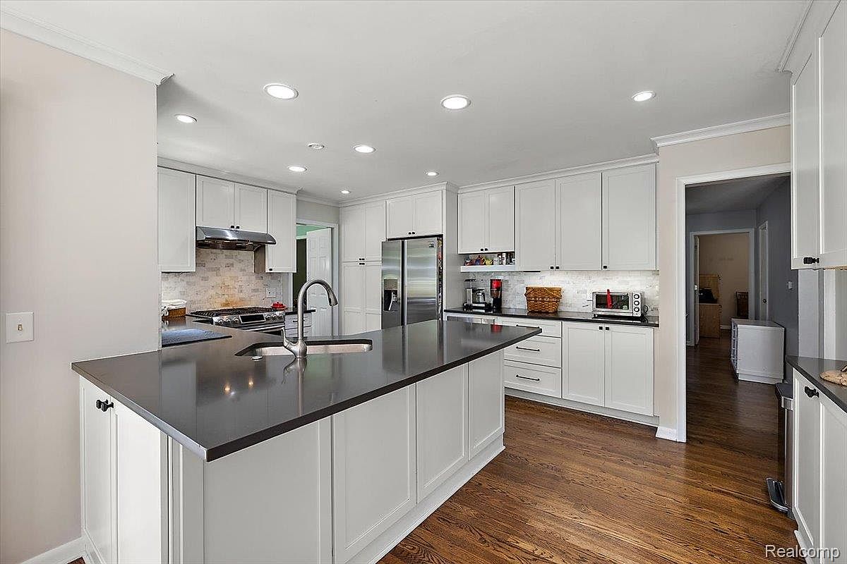 This is a bright and modern kitchen featuring white cabinetry, dark countertops, and stainless steel appliances. A large island with a sink provides ample workspace, and recessed lighting illuminates the area. The hardwood floors add warmth to the space, creating an inviting atmosphere.