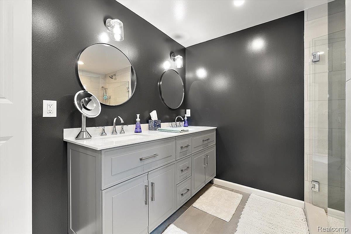 This is a primary bathroom featuring a double vanity with gray cabinetry and white countertops. Two round mirrors hang above the sinks, illuminated by modern sconces. The walls are painted a dark gray, and there is a glass-enclosed shower in the background, creating a modern and clean aesthetic.