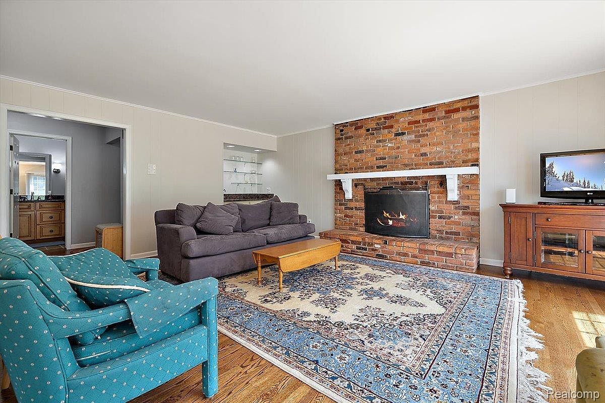 This is a cozy living room featuring a brick fireplace with a white mantel, a comfortable gray sofa, and a blue patterned armchair. A wooden coffee table sits on a large area rug with blue and beige patterns, adding warmth to the hardwood floors. A wooden TV cabinet stands against the wall, completing the inviting and traditional aesthetic.