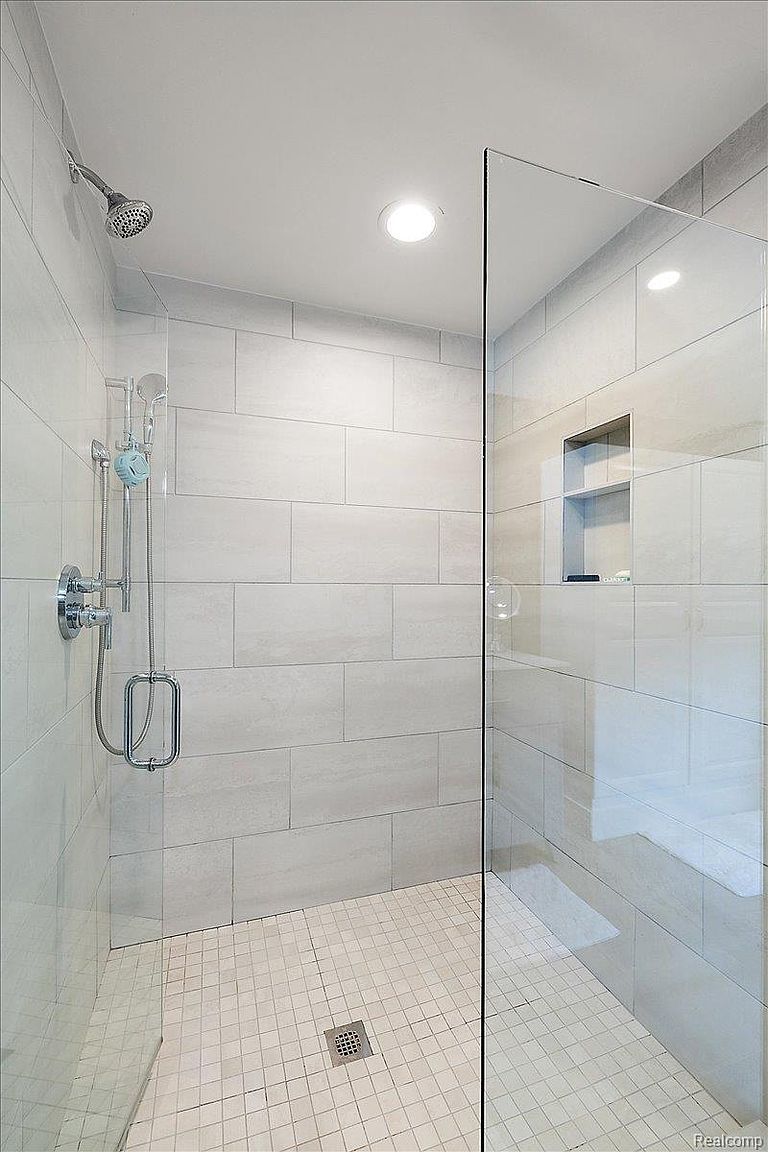 This is a well-lit primary bathroom featuring a modern shower with large, light-colored rectangular tiles on the walls and smaller square tiles on the floor. The shower has a glass enclosure, a built-in niche for storage, and chrome fixtures. The overall impression is clean, bright, and contemporary.