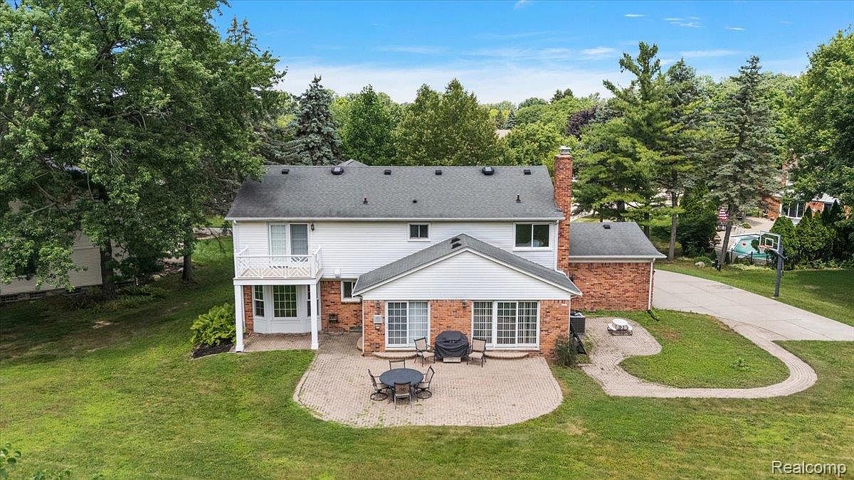 This is a rear view of a two-story house with a brick and white siding exterior. The house features a patio with outdoor furniture, a well-maintained lawn, and mature trees in the background. A brick chimney is visible, and the overall impression is one of a spacious and private backyard.