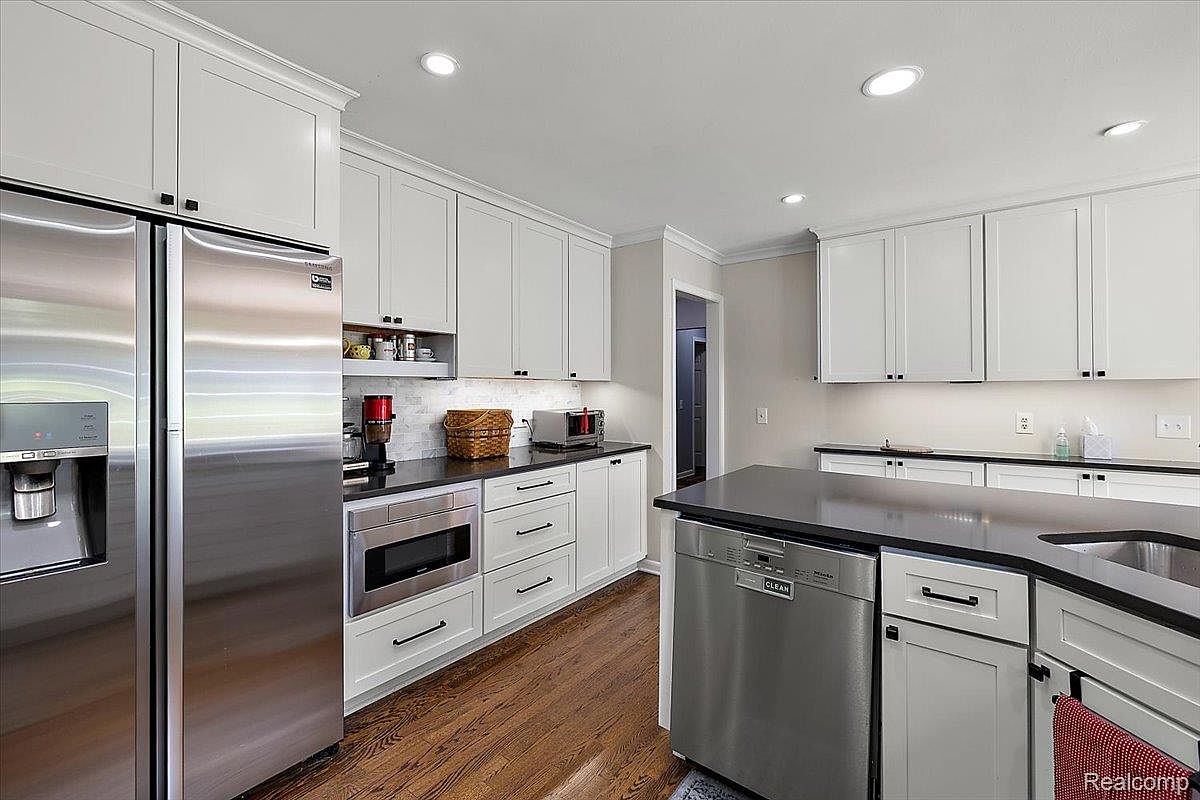 This is a well-lit kitchen featuring white cabinetry with dark hardware, stainless steel appliances, and dark countertops. The kitchen has hardwood flooring and recessed lighting. The perspective is from a corner, showcasing the refrigerator, microwave, dishwasher, and countertop space.