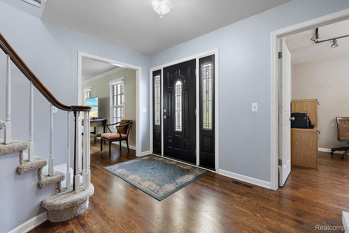 This is an interior shot of a home's entryway and hallway, featuring hardwood floors and light blue walls. A staircase with carpeted steps is visible on the left, while a black front door with decorative glass panels is centered. An open doorway leads to another room, suggesting a spacious and welcoming layout.