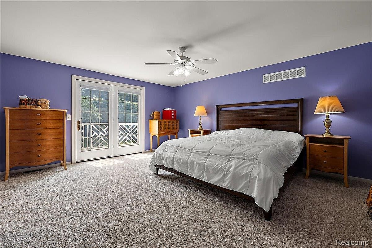 This primary bedroom features a calming purple color scheme, complemented by neutral carpeting. A large bed with a white comforter is the focal point, flanked by matching nightstands and lamps. French doors provide access to the outside, and a ceiling fan adds to the room's comfort.