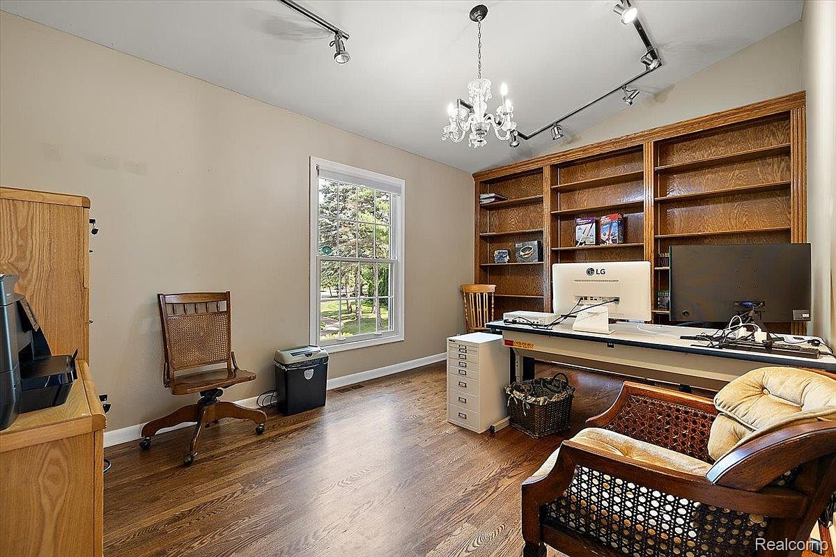This is an interior shot of a home office or study. The room features hardwood floors, built-in bookshelves, a desk with computer monitors, and a window providing natural light. There are two chairs in the room, one a rolling desk chair and the other an upholstered armchair, suggesting a comfortable and functional workspace.