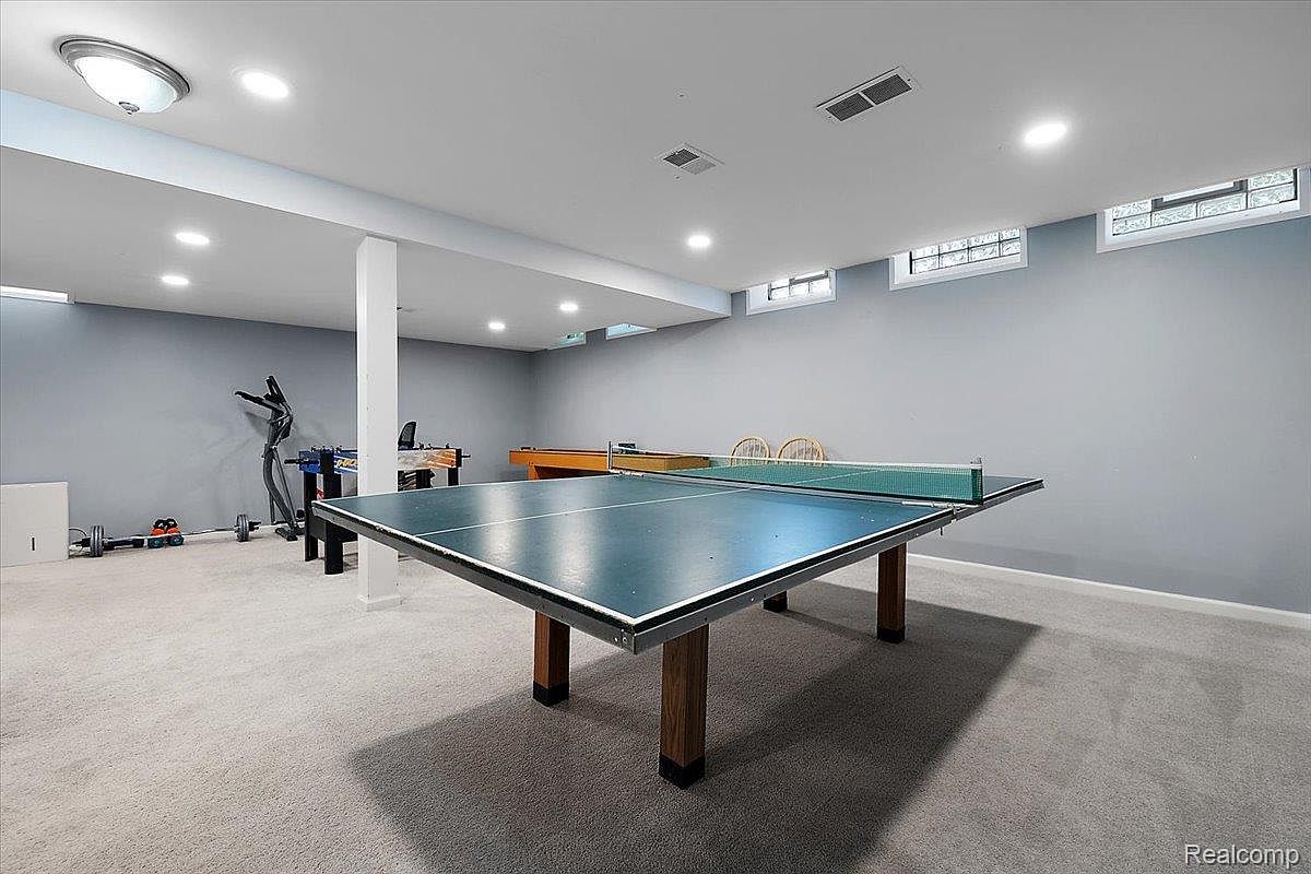 This is a spacious basement featuring a ping pong table as a central element. The room is well-lit with recessed lighting and has a neutral gray color scheme. Other features include a foosball table, exercise equipment, and small glass block windows near the ceiling, suggesting a functional and recreational space.