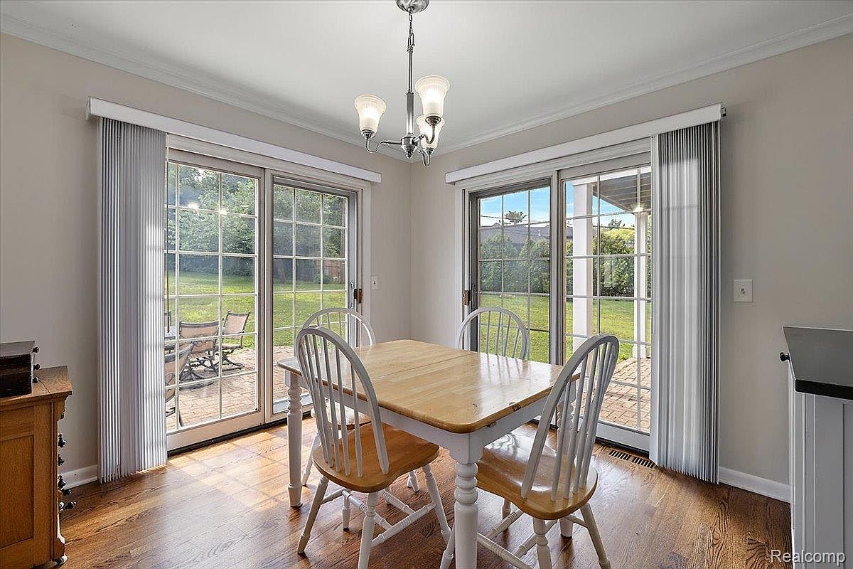 This is a bright and airy dining room featuring a wooden table with white legs and matching chairs. Natural light floods the space through two sets of sliding glass doors, offering views of the outdoor greenery. The room is simply decorated, creating a welcoming and comfortable atmosphere.