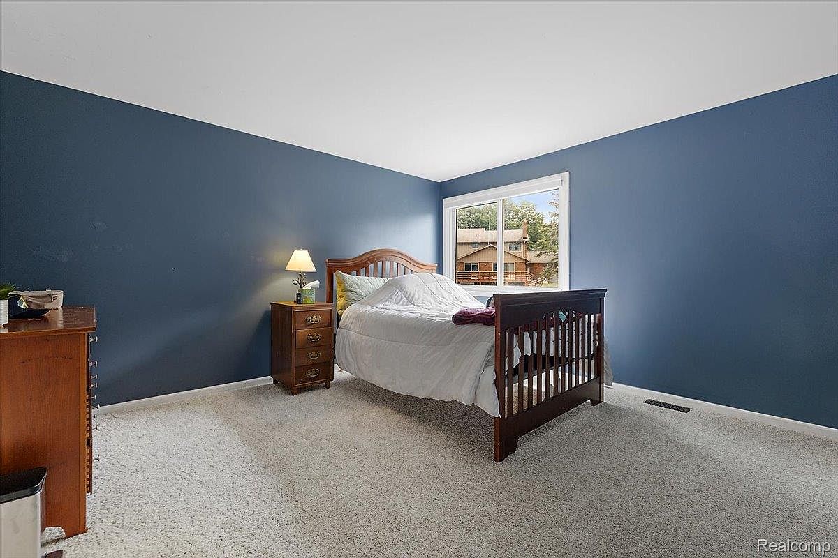 This is a cozy bedroom featuring a wooden bed frame with white bedding and a burgundy throw. The walls are painted a calming blue, complemented by light gray carpeting. A wooden nightstand with a lamp sits beside the bed, and a window provides natural light, creating a serene and inviting atmosphere.