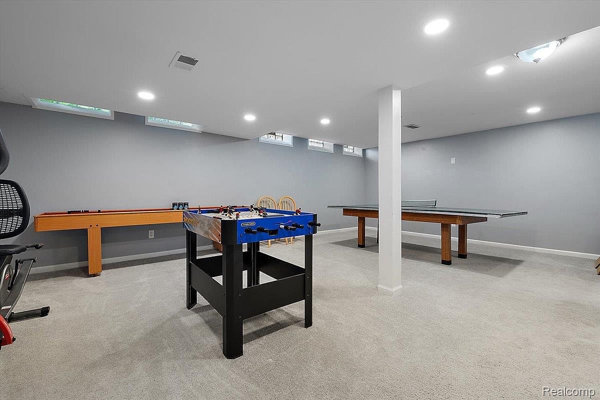 This is a spacious basement featuring a foosball table, a ping pong table, and a shuffleboard table, suggesting a recreational area. The walls are painted in a neutral gray tone, and the flooring is carpeted, creating a comfortable and inviting atmosphere. Recessed lighting provides ample illumination throughout the room.