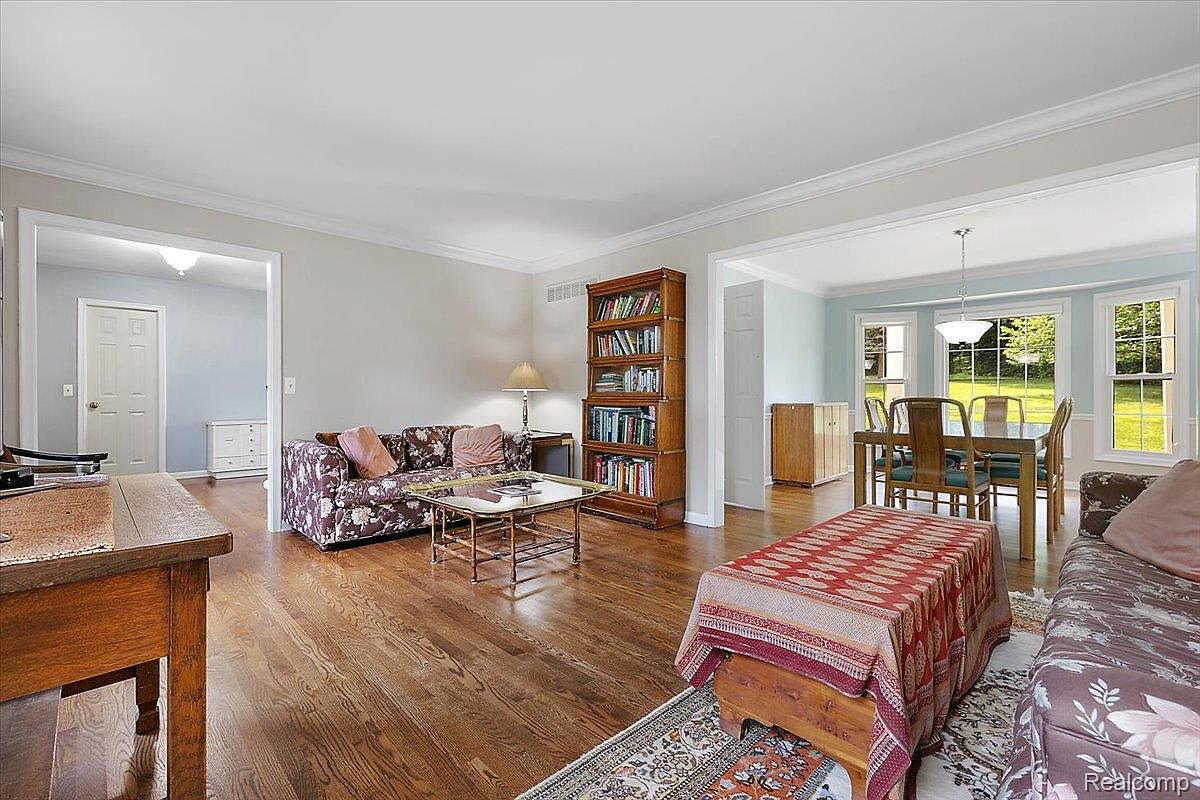 This is an interior shot of a living room featuring hardwood floors, a floral patterned sofa, and a wooden bookcase filled with books. The room opens into a dining area with a table and chairs, and natural light streams in through the windows. The overall impression is warm and inviting.