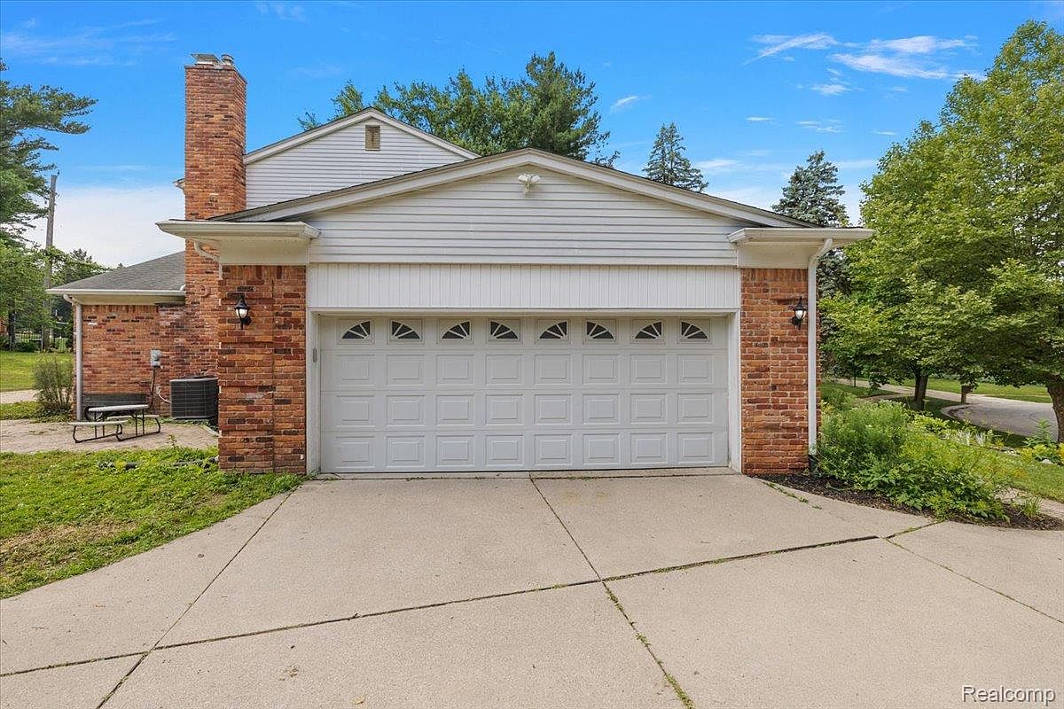 The image showcases a two-car garage with a white door featuring small, triangular windows along the top. The garage is flanked by brick walls and has a white, gabled roof. A concrete driveway leads up to the garage, and landscaping is visible on either side. The overall impression is of a well-maintained and functional garage space.