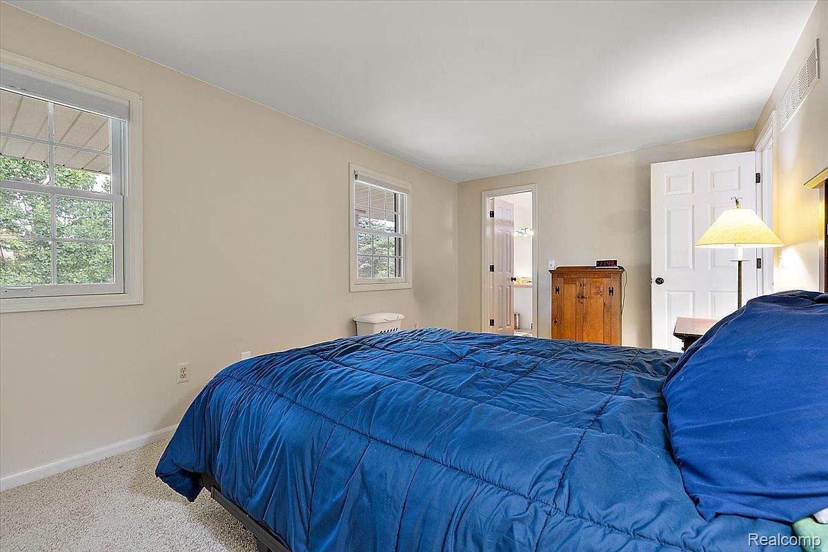 This is a cozy bedroom featuring a blue quilted bedspread, neutral-colored walls, and natural light streaming through two windows. A wooden cabinet stands against the wall, and a white door leads to another room. The room has a simple and comfortable aesthetic, suitable for a guest bedroom.