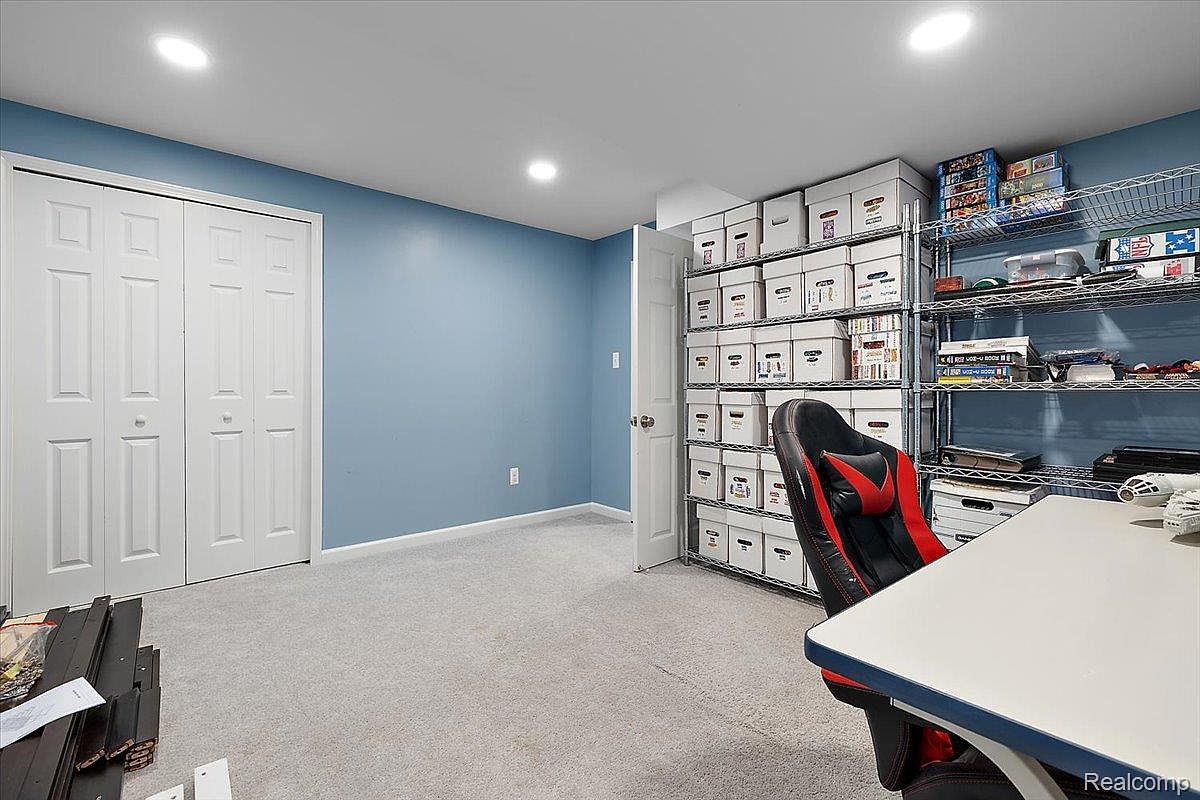 This is a well-organized basement space, likely used as a home office or storage area. The room features light blue walls, light gray carpet, and a double door closet. Shelving units filled with storage boxes and other items line one wall, and a desk with a gaming chair is positioned in the foreground. The room is illuminated by recessed lighting.