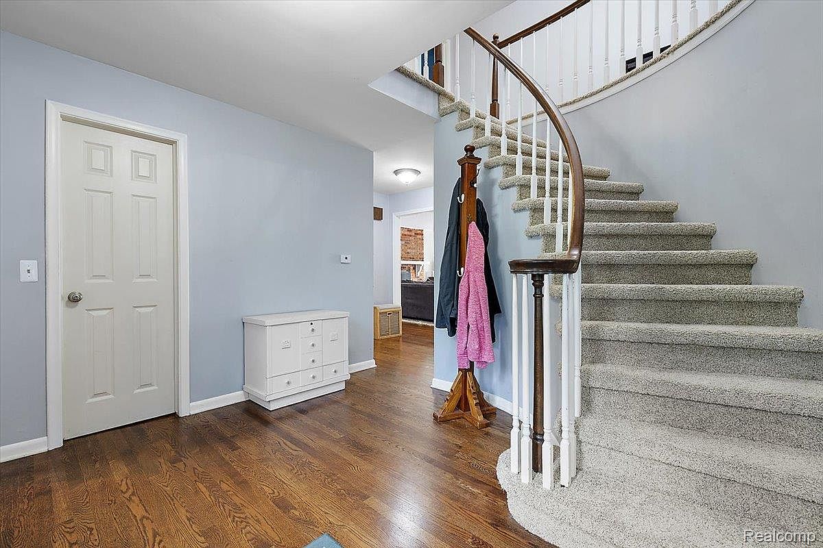 This interior shot showcases a welcoming hallway featuring a curved staircase with carpeted steps and a white railing with a wooden handrail. The walls are painted a soft blue, complemented by hardwood flooring and a white storage cabinet. A coat rack with hanging garments adds a touch of homeliness to the space.