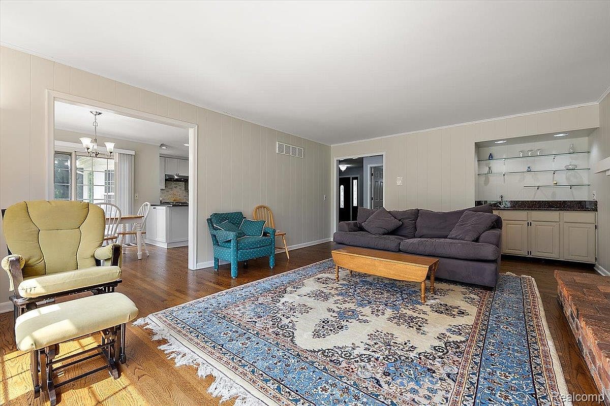 This is an interior shot of a living room featuring a large area rug with blue patterns, a gray sofa, and a wooden coffee table. There is a green rocking chair on the left side of the image and a built-in bar area with glass shelves on the right. The room has a cozy and inviting atmosphere, with hardwood floors and neutral-colored walls.