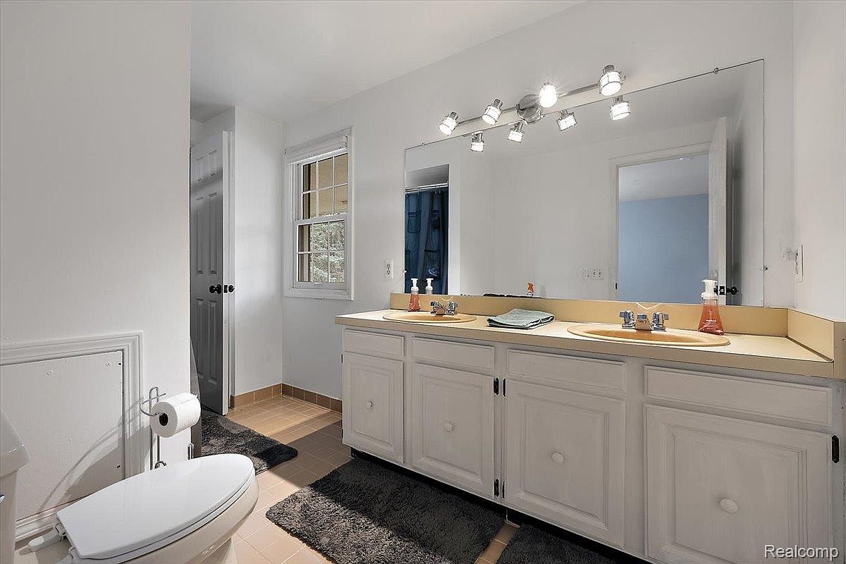 This is a well-lit bathroom featuring a double vanity with white cabinets and a light-colored countertop. A large mirror spans the length of the vanity, reflecting the bright overhead lighting. The room includes a toilet and a window, contributing to a clean and functional space.