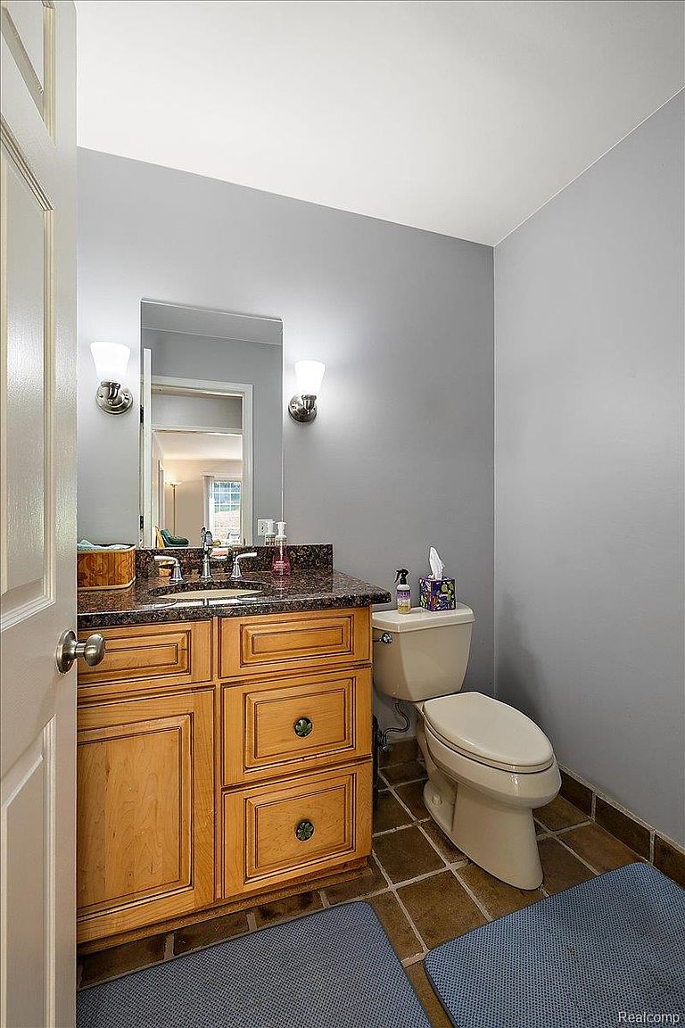 This is a cozy guest bathroom featuring a wooden vanity with a dark countertop, a rectangular mirror with sconces, and a standard toilet. The walls are painted a neutral gray, and the floor is tiled in a warm brown tone. Blue mats add a touch of color to the space, creating a welcoming and functional area.