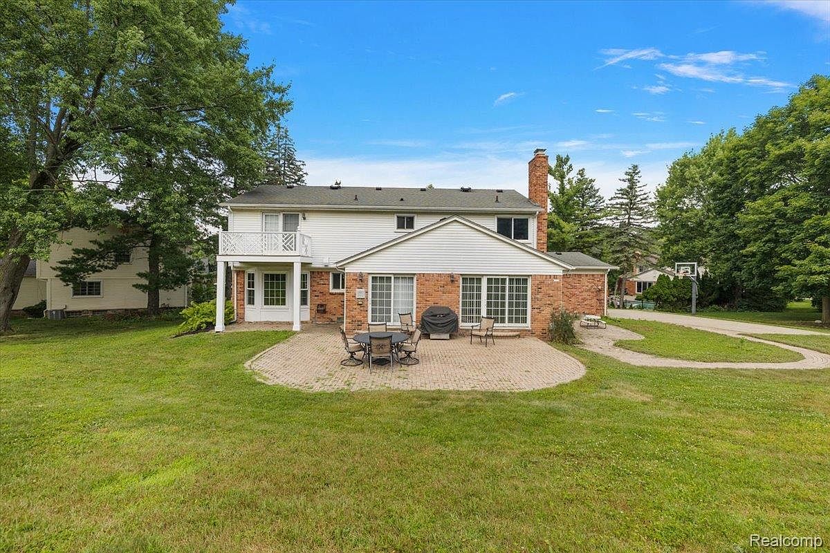 This is a rear view of a two-story house with a brick and white siding exterior. A brick patio with outdoor furniture is visible in the backyard, which is covered in green grass. The house features a balcony and a brick chimney, and is surrounded by mature trees.