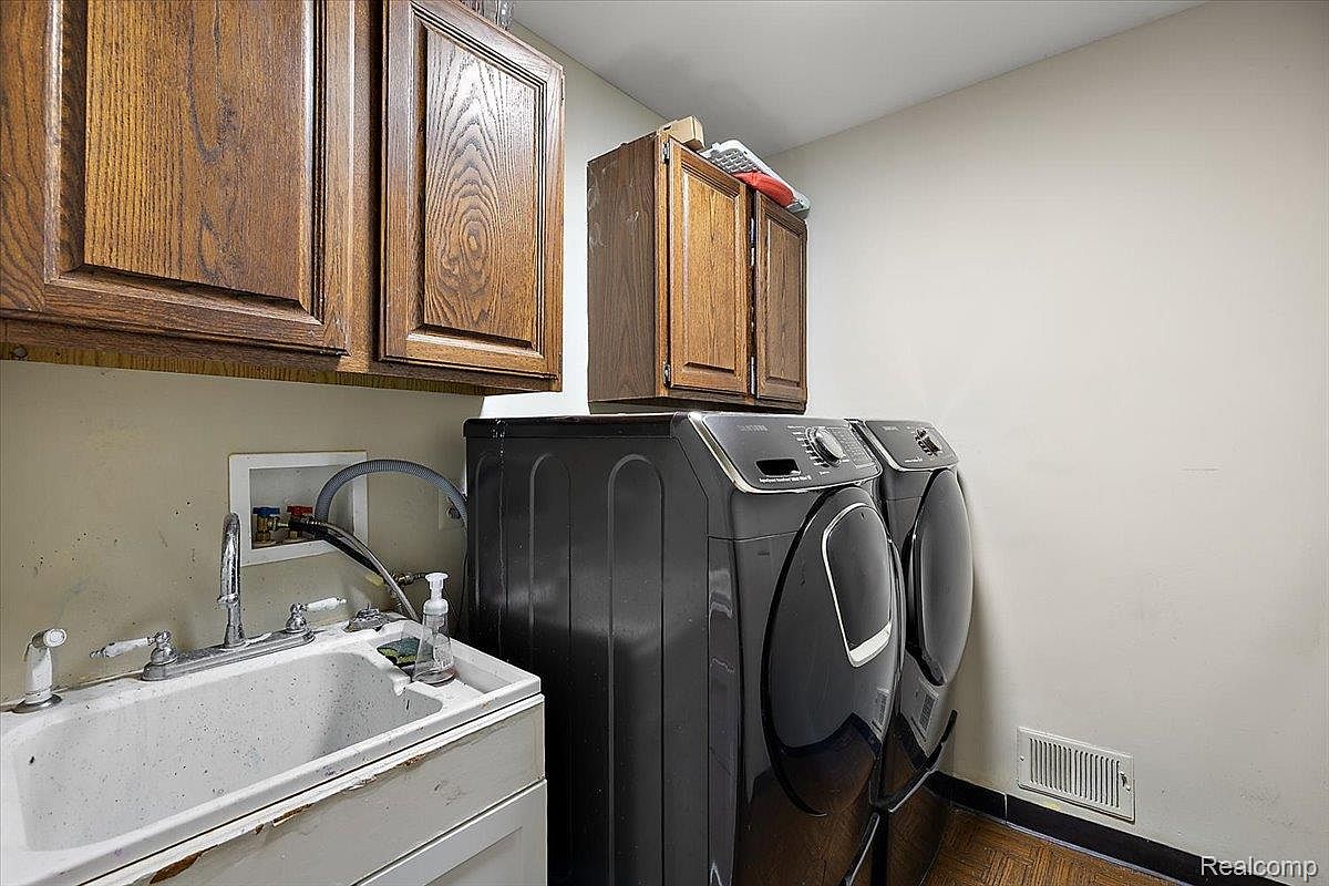 This laundry room features a utility sink with chrome fixtures, dark-colored front-loading washer and dryer set, and wooden cabinets above. The room has a functional and practical design, suitable for everyday laundry needs. The flooring appears to be wood or laminate.