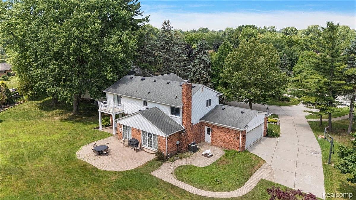 This aerial view showcases a well-maintained two-story home with a brick and siding exterior, complemented by a dark gray roof. The property features a spacious backyard with mature trees, a patio area with outdoor seating, and a long driveway leading to an attached garage. The overall impression is one of a private and inviting residence.