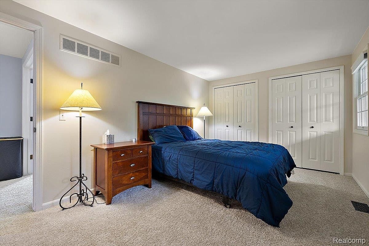 This is a primary bedroom featuring a wooden headboard bed with blue bedding, a wooden nightstand with a lamp, and two closet doors. The room has neutral-colored walls and carpet, creating a calm and inviting atmosphere. The perspective is from the doorway, showcasing the layout and key elements of the room.