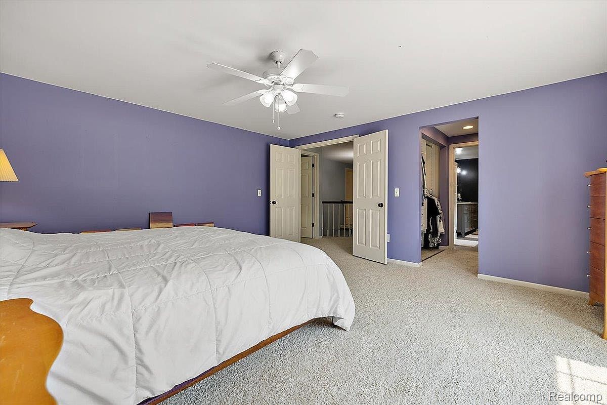 This is a primary bedroom featuring a large bed with a white comforter, light purple walls, and light-colored carpeting. A ceiling fan provides air circulation, and double doors lead to a hallway and a walk-in closet. The room has a calm and inviting atmosphere.