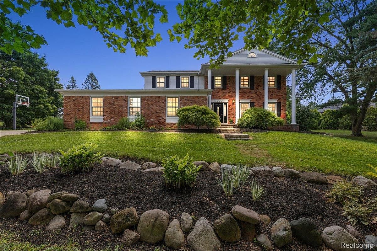 This is a front exterior view of a two-story brick house with white columns supporting a covered entryway. The house features symmetrical window placement with black shutters and a well-manicured lawn with a rock-lined garden bed in the foreground. The overall impression is one of classic elegance and curb appeal.