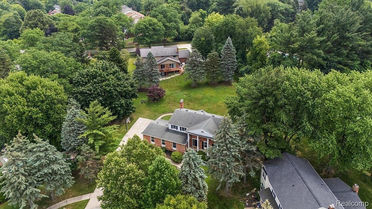 This aerial shot showcases a well-maintained property featuring a brick house with a dark roof, surrounded by lush greenery and mature trees. A second house is visible in the background, and the overall impression is one of privacy and spaciousness. The image highlights the property's setting within a verdant, established neighborhood.