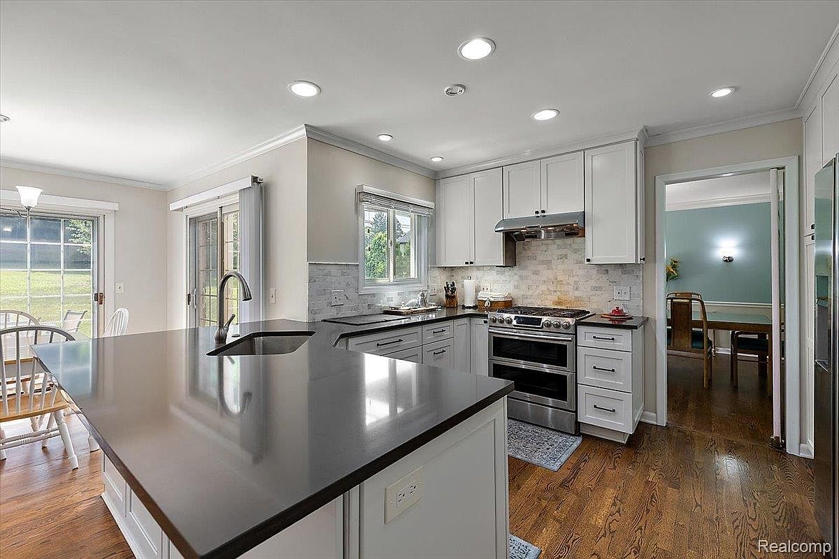 This is a well-lit kitchen featuring white cabinets, stainless steel appliances, and a dark countertop island with a sink. The kitchen has hardwood floors and a neutral color scheme, creating a clean and modern aesthetic. The view is from a wide angle, capturing the island, stove, and doorway to another room.