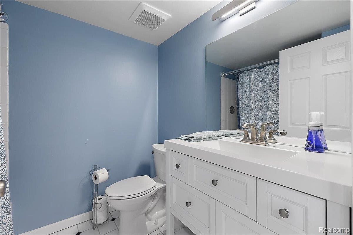 This is a well-lit bathroom featuring a blue wall, a white toilet, and a white vanity with a sink. A large mirror reflects the shower area with a patterned curtain. The overall impression is clean and functional, suitable for a guest bathroom.