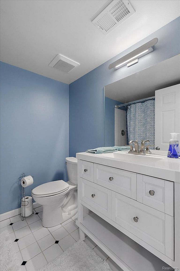 This is a bathroom featuring a white vanity with drawers and cabinets, a white toilet, and a mirror above the sink. The walls are painted a light blue, and the floor is tiled in white with black accents. The bathroom appears clean and well-maintained, offering a functional and pleasant space.