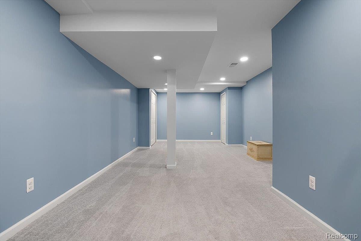 This is a spacious, finished basement with light blue walls and gray carpeting. The room is well-lit with recessed lighting and features a support column in the center. Two closet doors are visible, and a wooden chest sits in the corner, suggesting ample storage space.