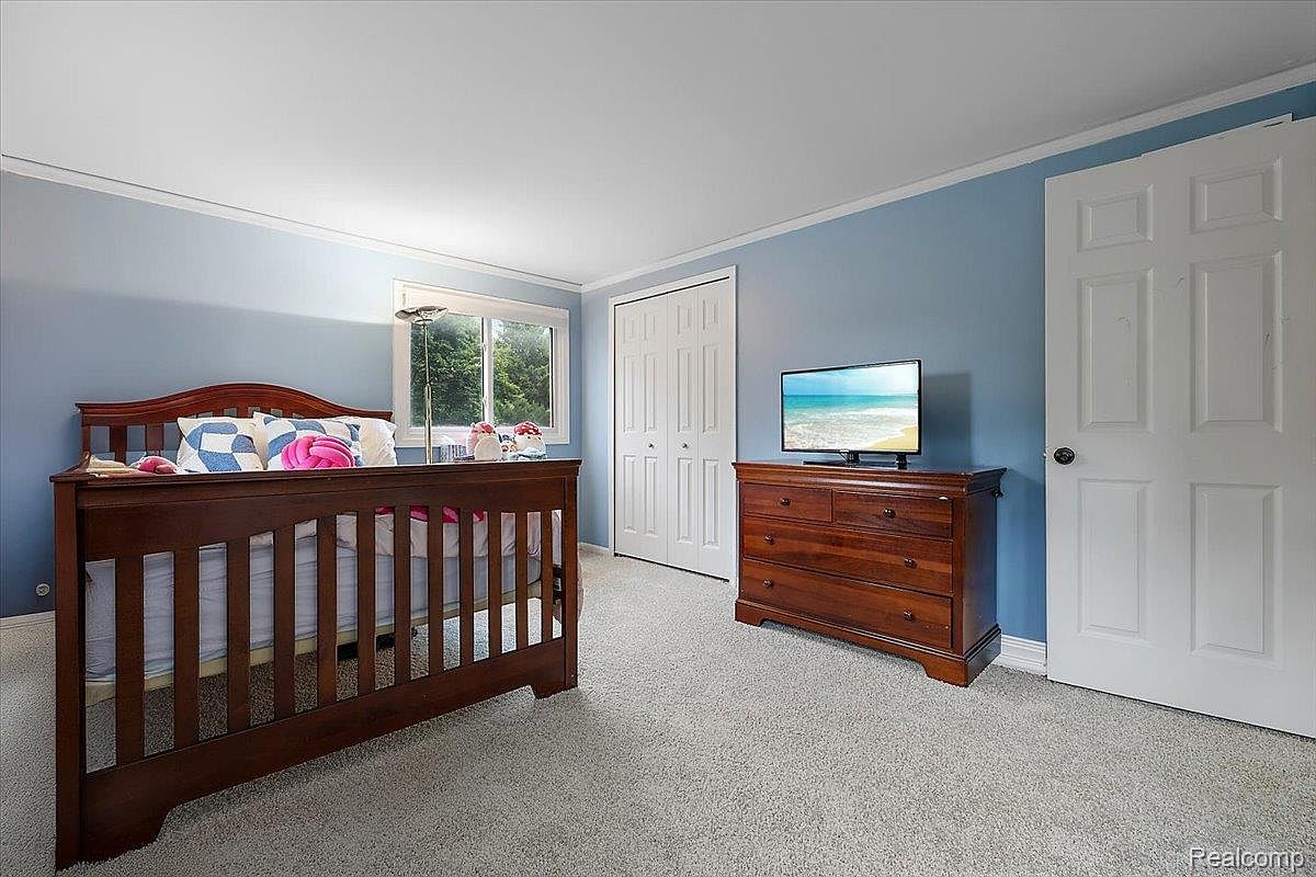 This is a cozy bedroom featuring a wooden crib with bedding, a wooden dresser with a television on top, and a white closet and door. The walls are painted a calming blue, and the room is carpeted in a neutral tone. The overall impression is a comfortable and inviting space.