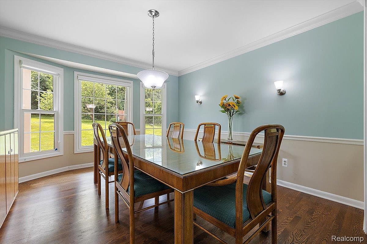 This is an interior shot of a dining room featuring a wooden dining table with a glass top and six chairs with green cushions. The room has a light blue upper wall and a beige lower wall, separated by white trim. Natural light streams in through multiple windows, and a chandelier hangs above the table, creating a bright and inviting atmosphere.