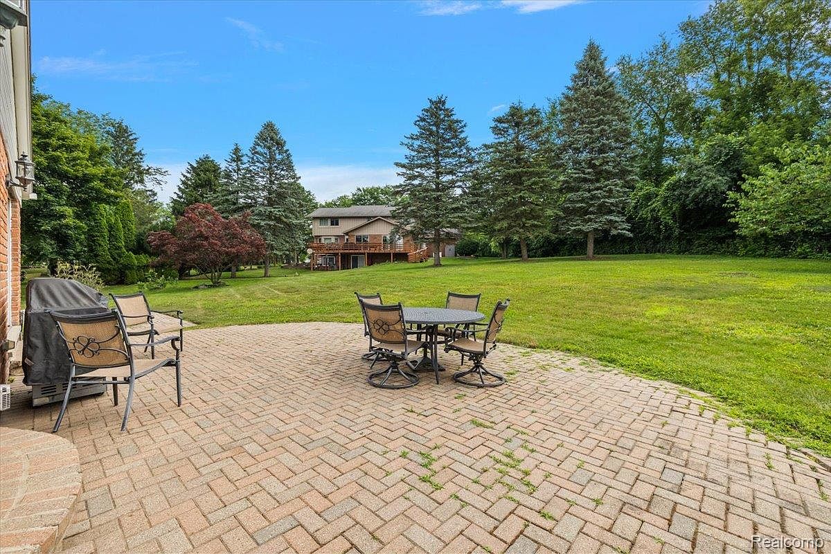 This image showcases a spacious brick patio with an outdoor dining set, including a round table and several chairs. The patio overlooks a well-maintained lawn and mature trees, with a house visible in the background. The scene conveys a sense of outdoor living and relaxation.