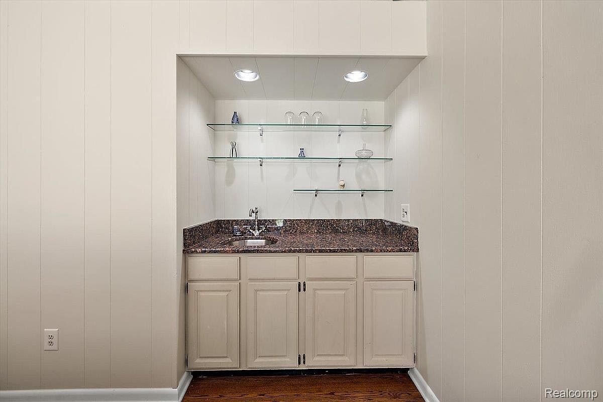 This interior shot showcases a built-in wet bar area, featuring a dark countertop, light-colored cabinetry, and glass shelving displaying various glassware. The recessed lighting highlights the setup, creating an inviting space for entertaining. The walls are paneled, adding a touch of traditional style.