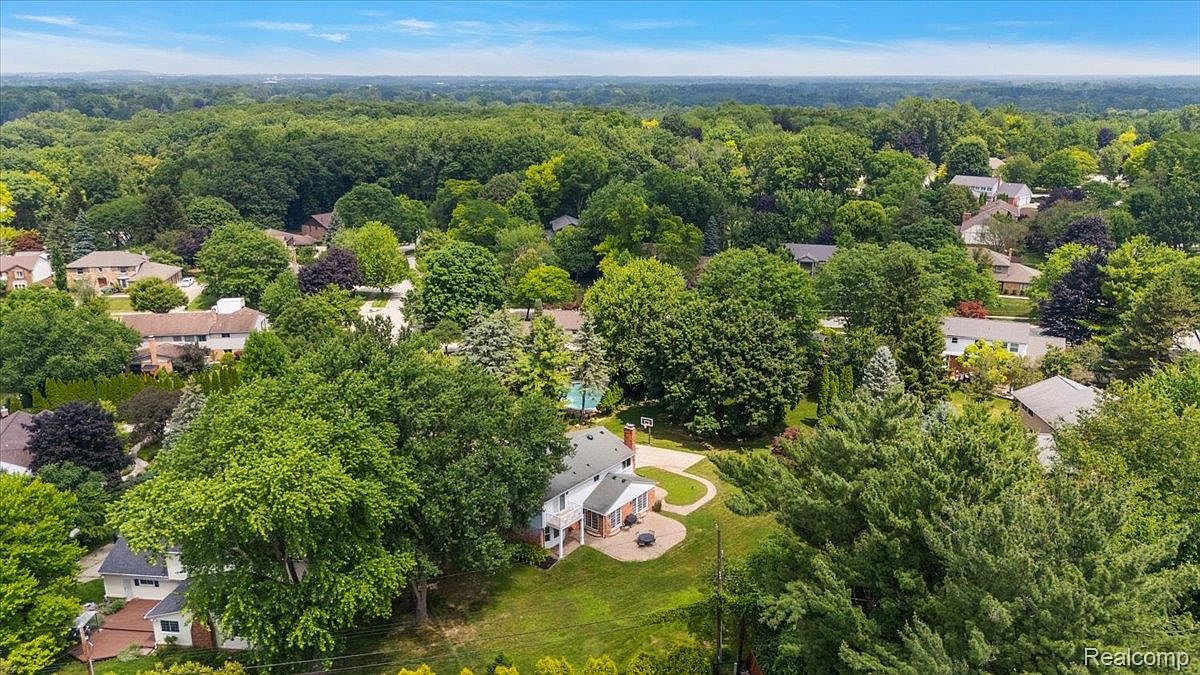 This aerial shot showcases a beautiful residential area with lush green trees and well-maintained homes. The featured property boasts a spacious backyard with a patio area, a basketball hoop, and a swimming pool, suggesting a focus on outdoor living and recreation. The overall impression is one of a peaceful and desirable neighborhood.