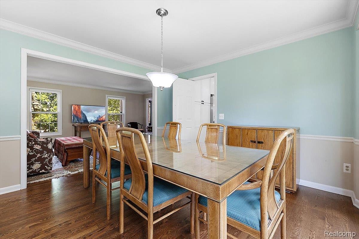This is an interior shot of a dining room featuring a glass-topped dining table with wooden chairs and blue cushions. A modern light fixture hangs above the table, and a wooden cabinet is positioned against the wall. The room has a light blue upper wall and a white lower wall, with hardwood flooring adding warmth to the space. The perspective is from a medium distance, capturing the entire dining set and a glimpse into an adjacent living area.