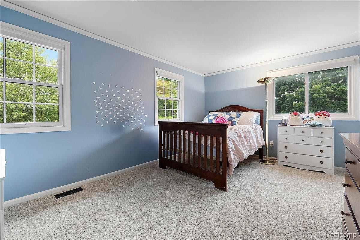 This is a cozy bedroom featuring light blue walls, white trim, and neutral carpet. A dark wood crib-style bed is neatly made with a light pink comforter and decorative pillows. Natural light streams in through two windows, and a white dresser with decorative items sits against the wall, creating a warm and inviting atmosphere.